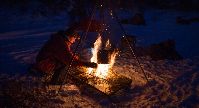 a person tends to a campfire in the snow. There is a tripod holding a pot over the fire. 